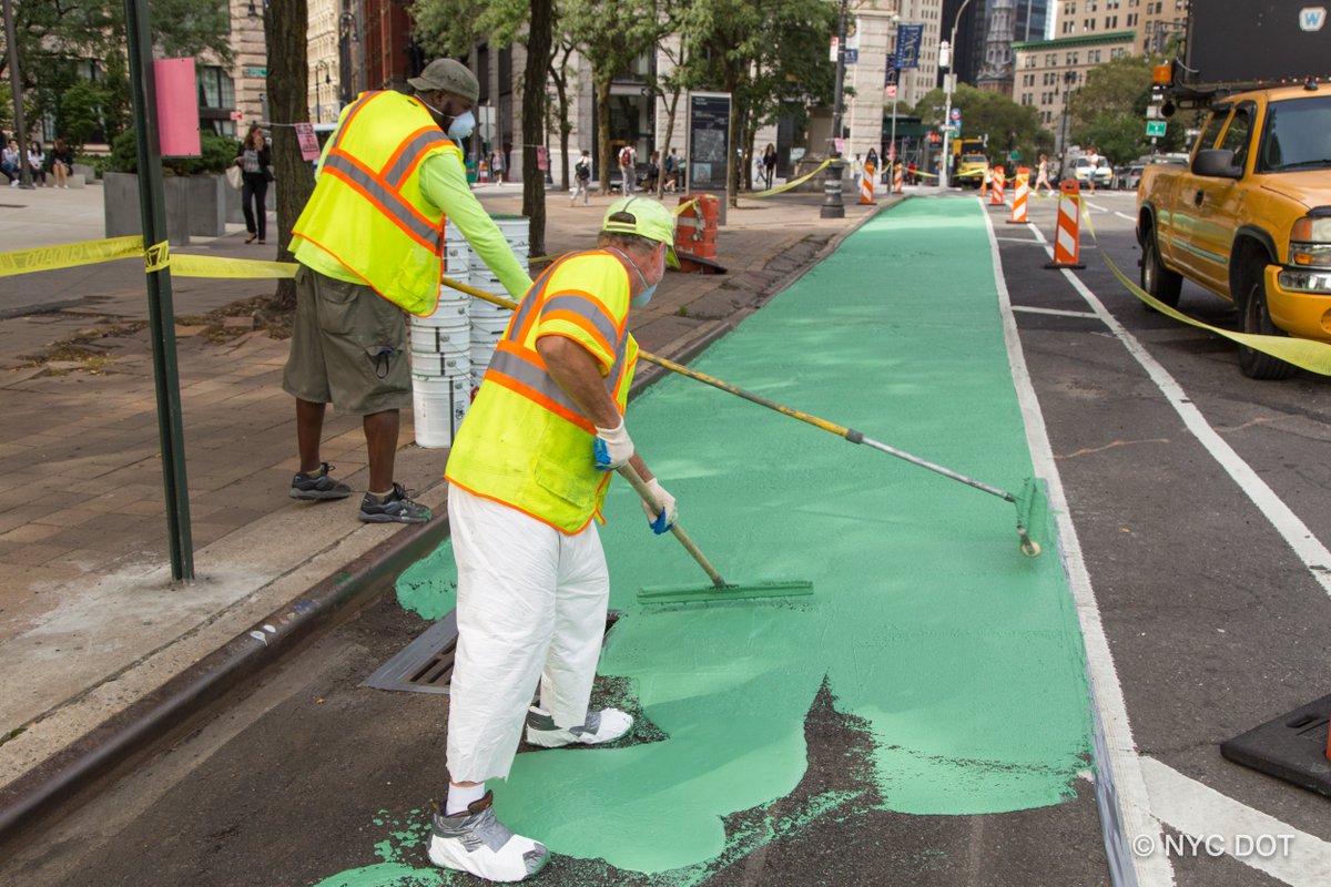 People in yellow and orange safety gear paint green paint on the roadway using rollers. 