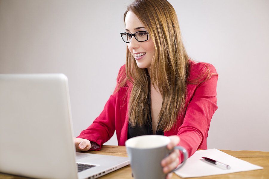 a woman drinking coffee at her desk smiling at the laptop in front of her