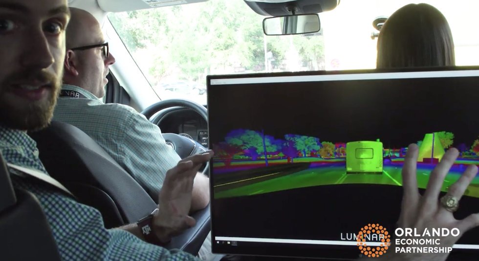 Three people are sitting in a vehicle. A young, bearded man (Nico)  is gesturing at a computer monitor that has a LiDAR-generated image of the road and vehicles ahead of them. The photo is credited to Orlando Economic Partnership.