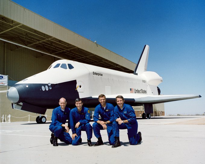 Four men in blue flight suits kneel in front of space shuttle