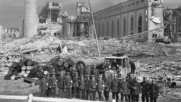 black and white photo of uniformed men standing in a line before two dozen stacked coffins with the ruins of a factory explosion in the background. 