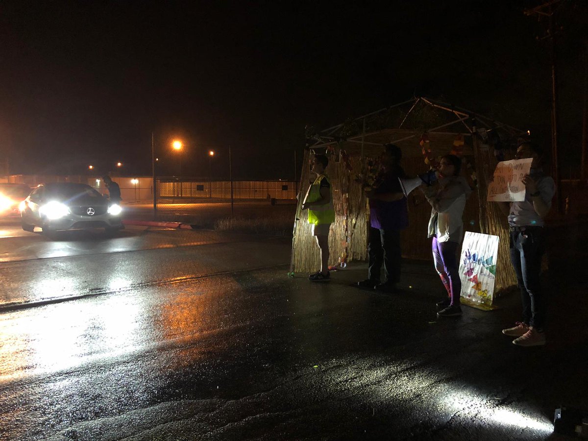A car tries to exit a detention centers. Protesters and a sukkah stand in it's way.