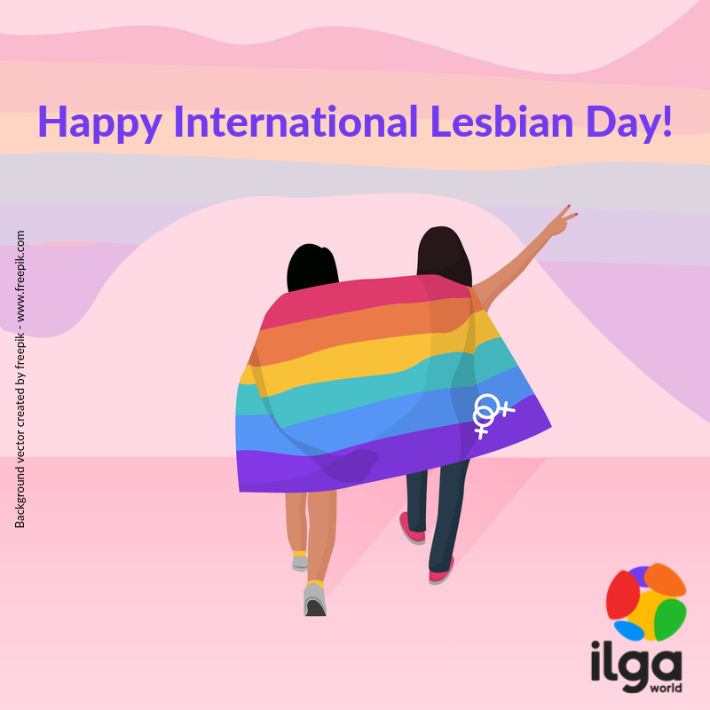 Two women hug below a rainbow flag. The image reads "Happy International Lesbian Day", and shows the ILGA World logo