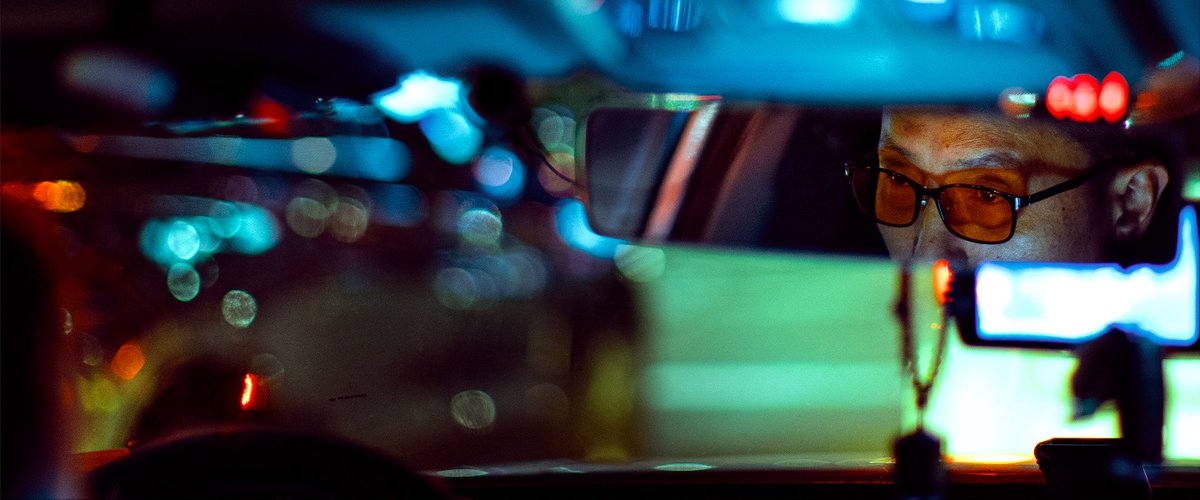 Photography by Liam Wong of Seoul at night. The interior of a taxi, with the mirror focused on the eyes of the driver. It is blue in color and is surrounded by bokeh from the raindrops and lights.