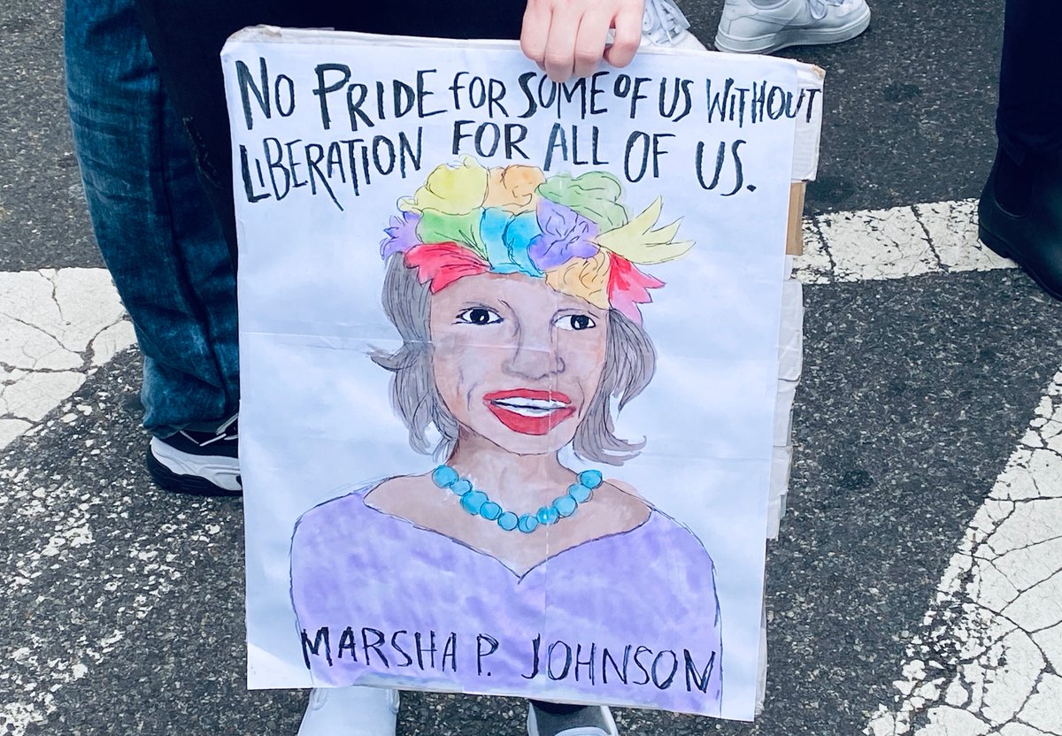 A protest sign contains an image of Marsha P. Johnson with the words "No pride for some of us without liberation for all of us" displayed at the top.