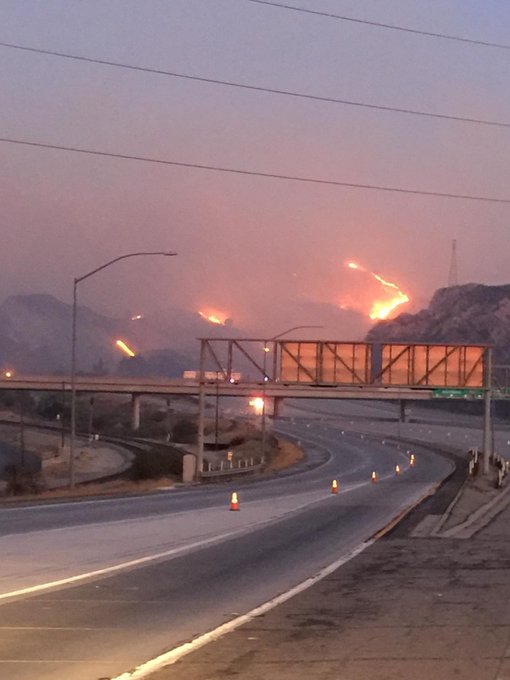 Fire in the hills near a closed freeway interchange where there are no cars and some traffic cones in lanes