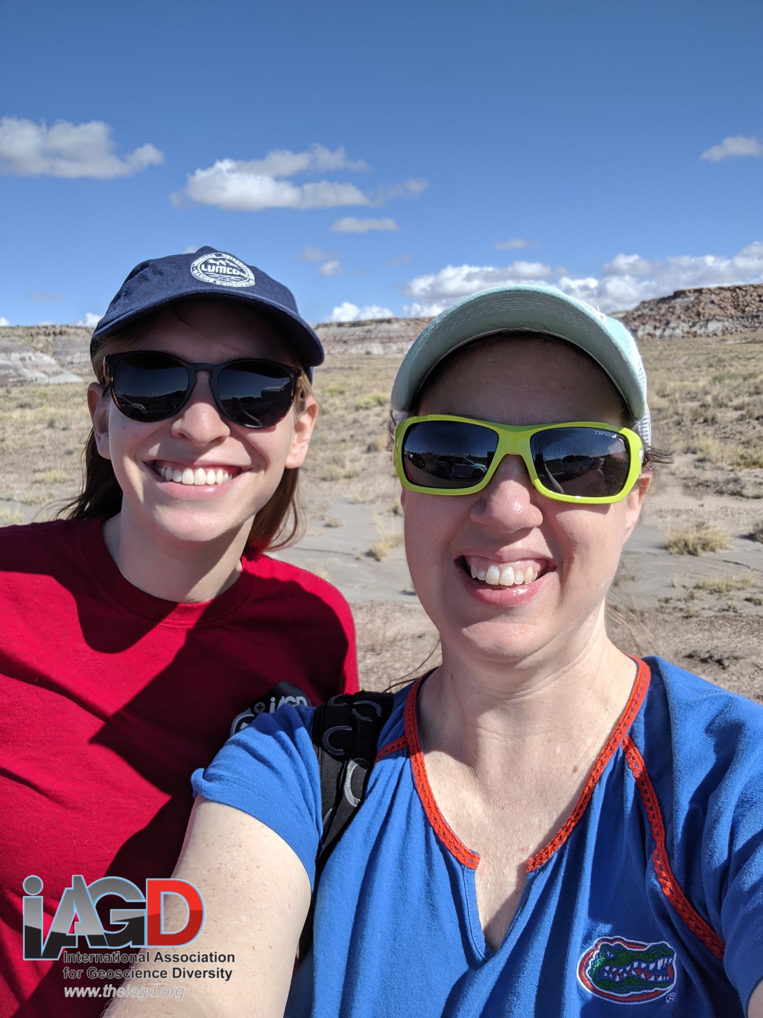 two women wearing sunglasses and hats are smiling. they're outside in the desert.