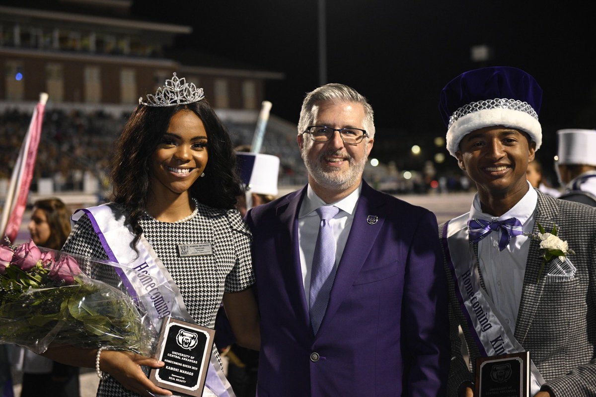 The homecoming king and queen in their crowns with President Davis in a purple suit on the football field.