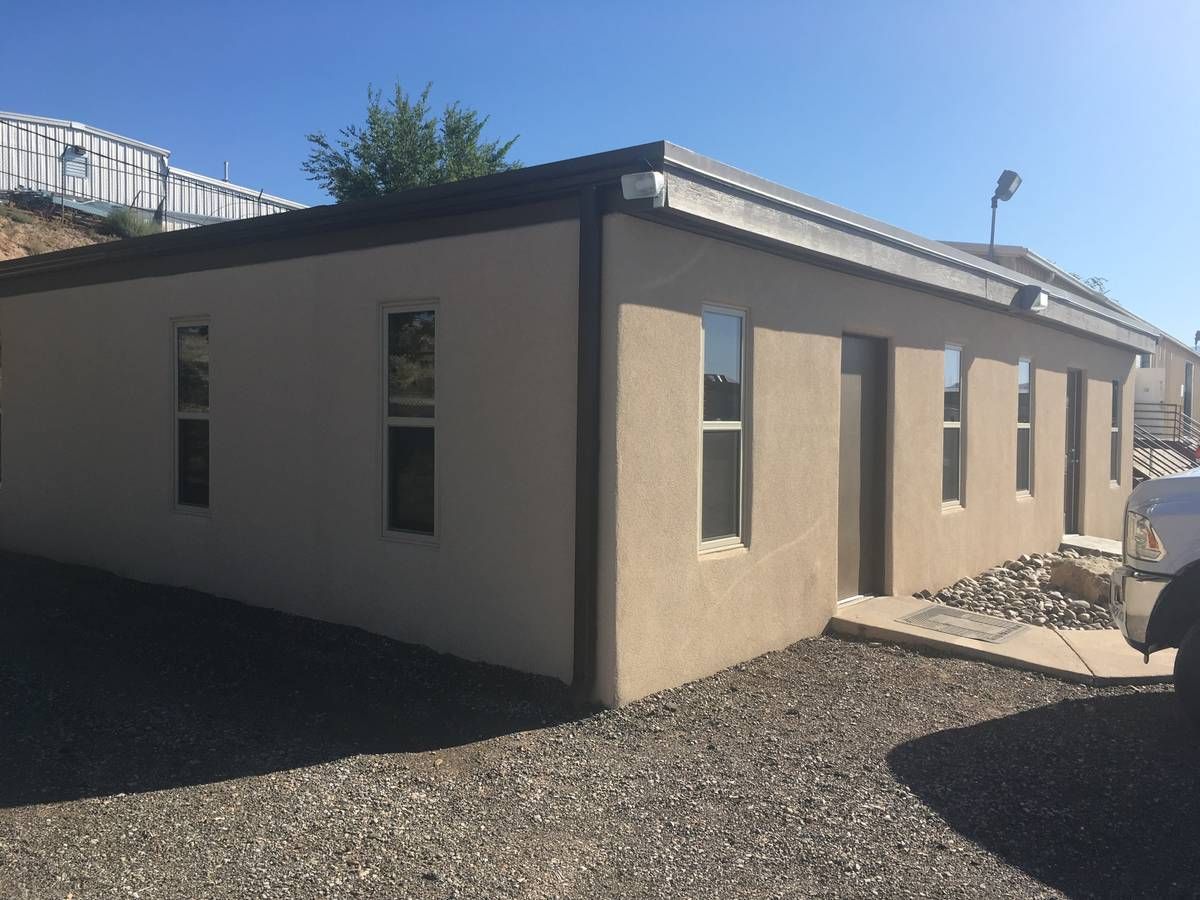 A photo of a small office building with a beige stucco facade surrounded by gravel beds. The front of a parked truck is visible on the right. In the background on the left, there is a vinyl-sided building at the top of a hill. in the background on the right, another small office building is visible.