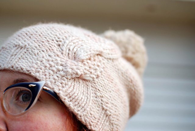 close up photo of a woman wearing a chunky knitted hat in a pale pinky/beige. Only a quarter of her face is visible and the background is blurred. The light suggests it was taken outdoors.