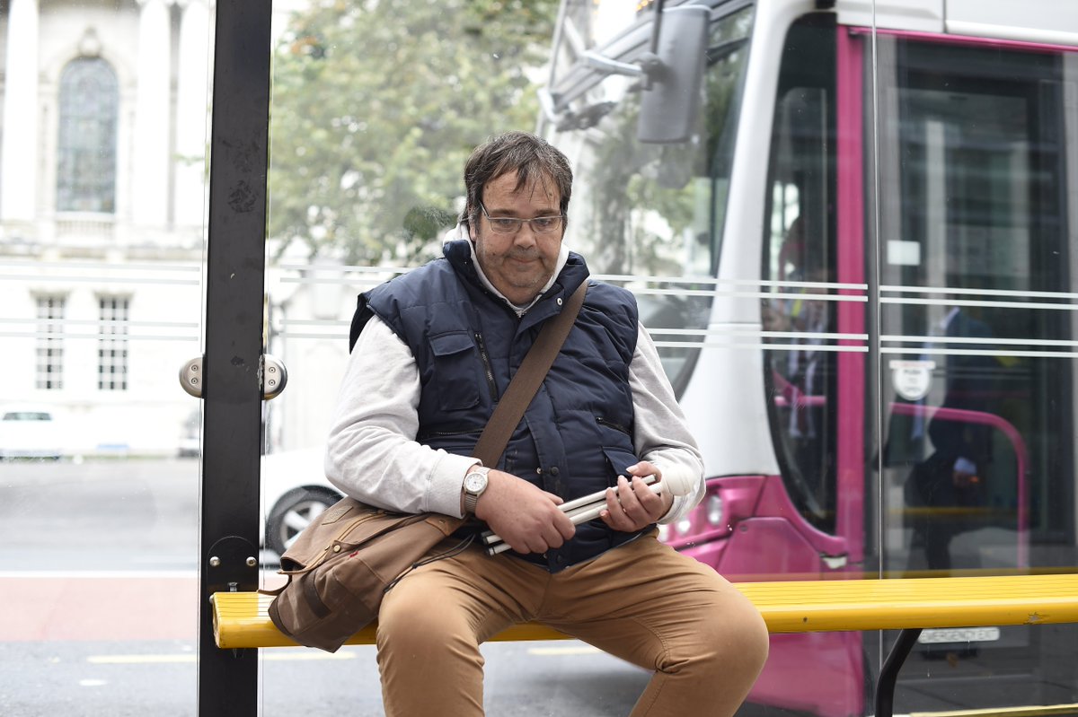 Image of man sitting at bus stop holding folded cane with bus in background