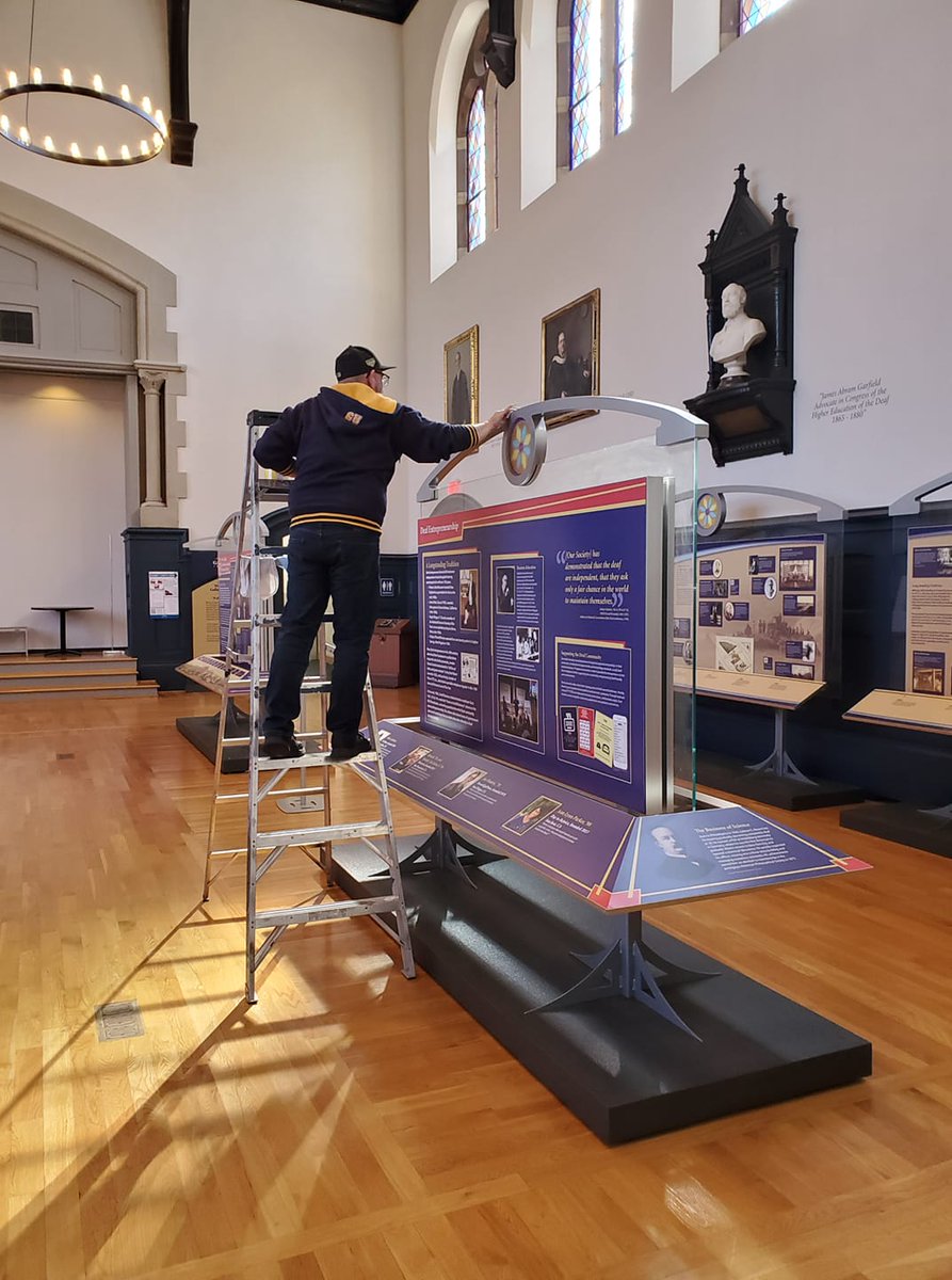 A man in a Gallaudet hoodie stands on a ladder cleaning a museum exhibit.