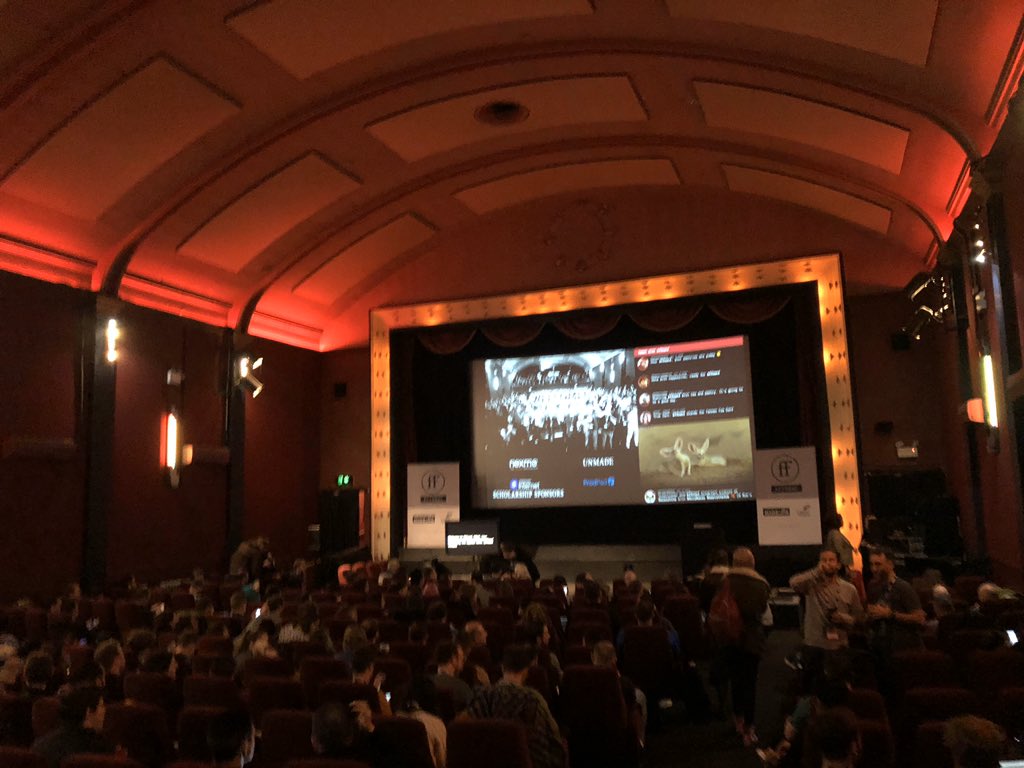 Inside the of Duke of York Picturehouse cinema, glowing pink/red ceiling above a cinematic screen bordered in a soft yellow glow. Filled with people attending FFConf.