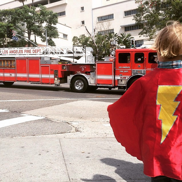 Child standing curbside wearing a superhero cape as a Los Angeles Fire Department truck rushes by.