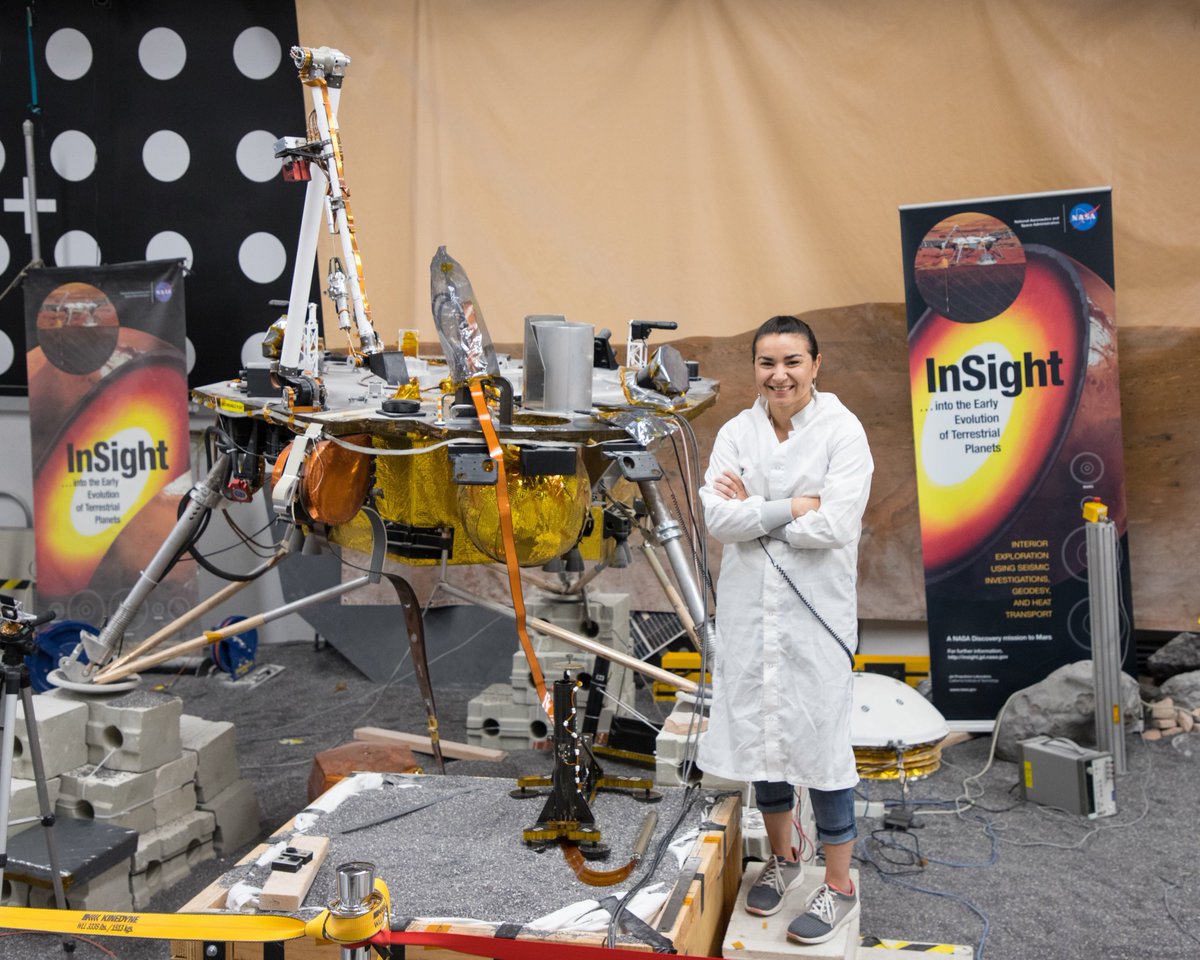 Marleen Sundgaard stands in a gravel sandbox with a test model of the InSight Mars lander behind her and InSight banners on either side of her. 