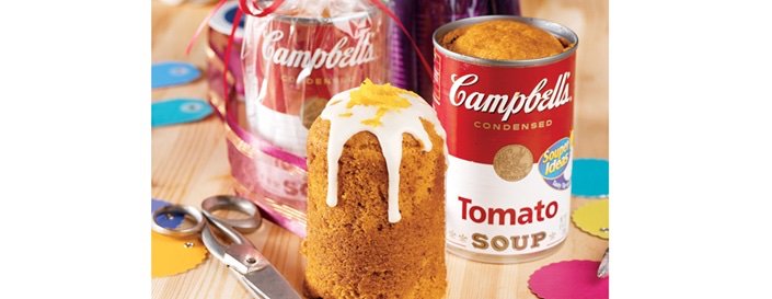 An orange, tall piece of tomato soup cake, frosted with a small amount of white frosting, beside two Campbell's Tomato Soup cans. From Campbell Soup's website.