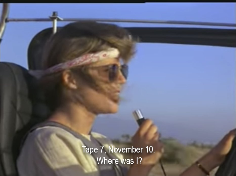 A woman wearing a headband and sunglasses, driving in an open-top car. She is holding a recorder microphone. The subtitle reads, "Tape 7, November 10. Where was I?"