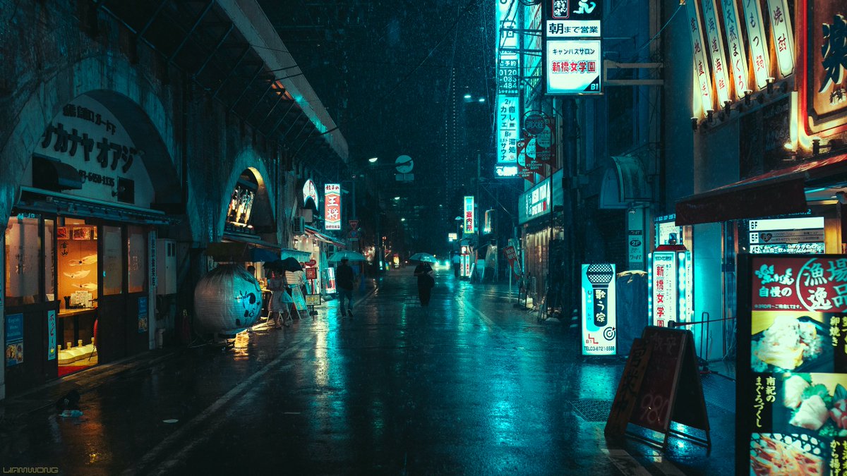 Photography by Liam Wong of Tokyo at night captured during a typhoon. The silhouette of a man carrying an umbrella walking towards the camera in the distance. It is teal and blue in color with red highlights. It is along a large overhead train station with restaurants under the arch.