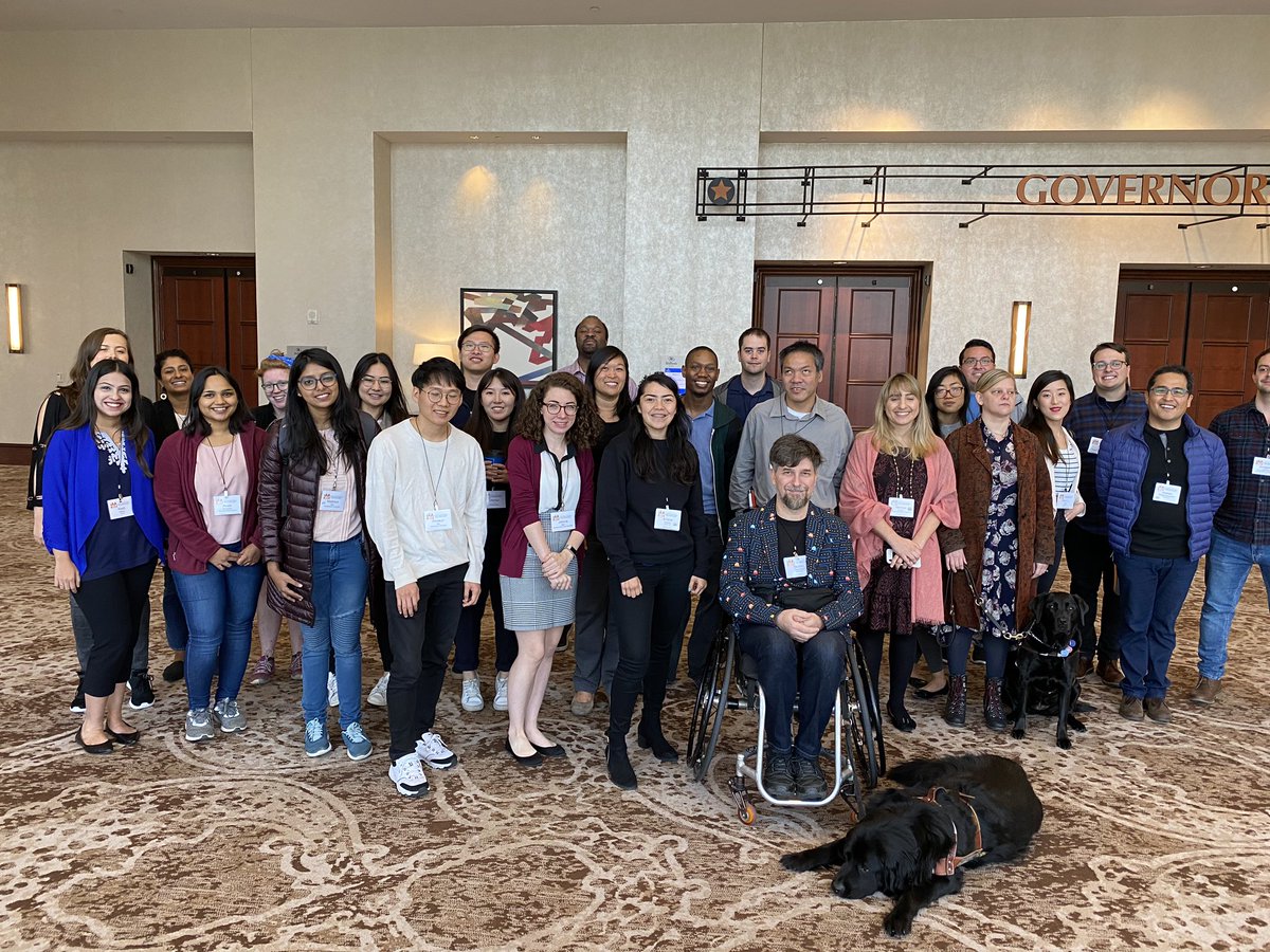 A group of around 25 people with two service dogs posing for a group photo in a hotel convention center.