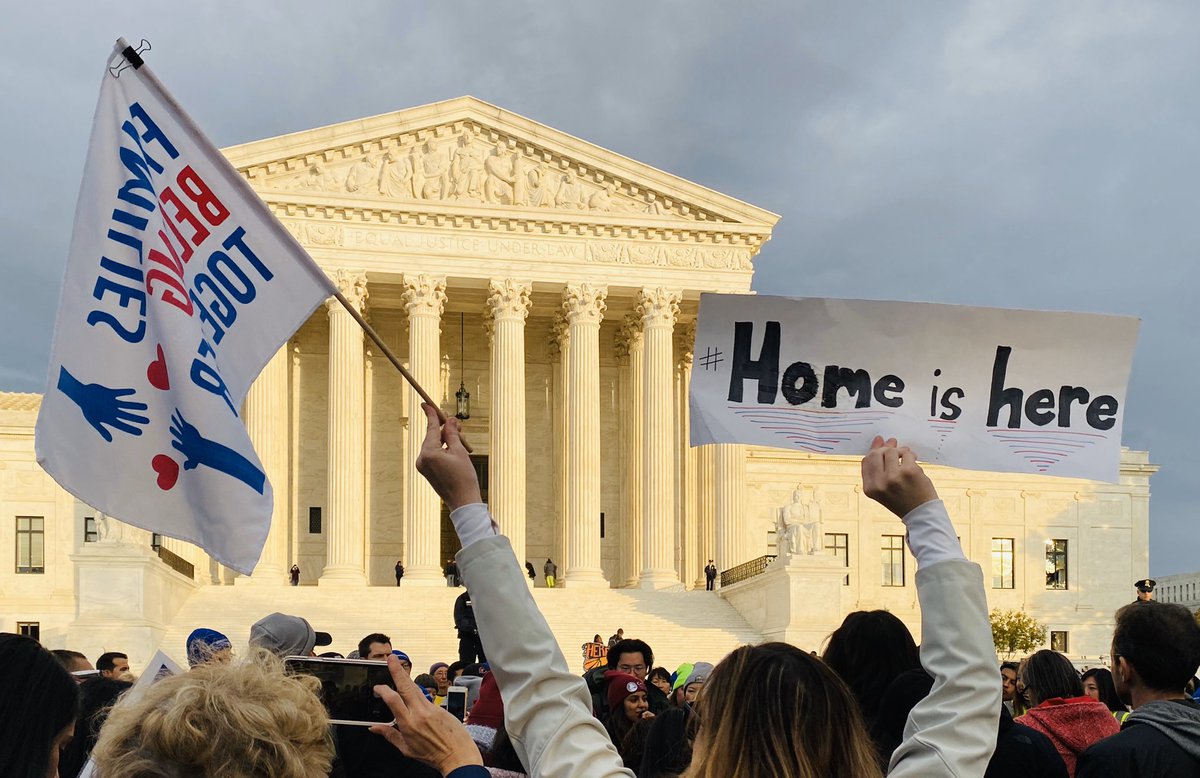 Woman in front of the Supreme Court holds a flag that says “families belong together” and a sign that says “home is here.”