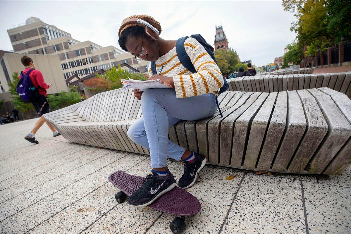 A student sits on a wooden bench, writing in a notebook while she rests her foot on her skateboard 