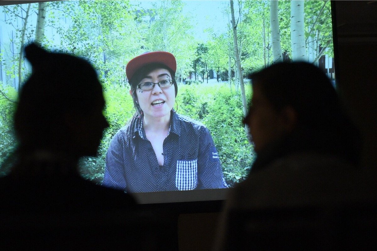 Two people watch a video of another person speaking in a forest.