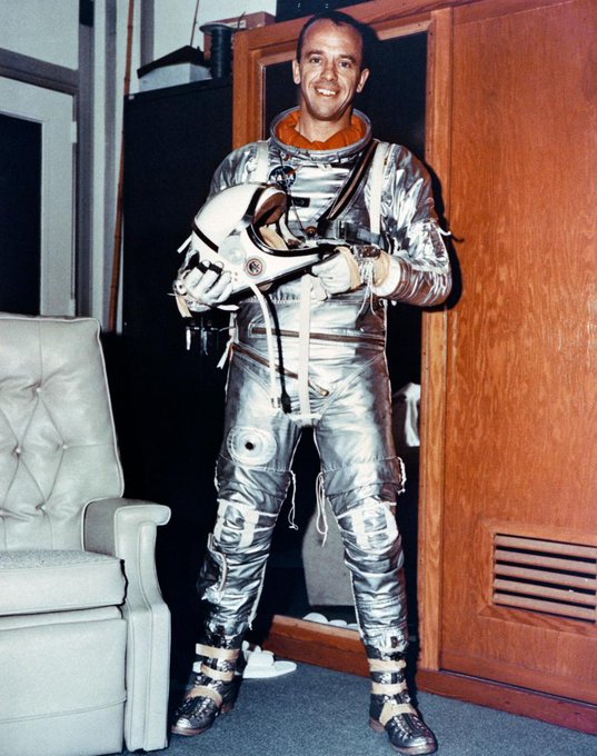 Alan Shepard stands in front of a wall locker in his silver Mercury spacesuit.
