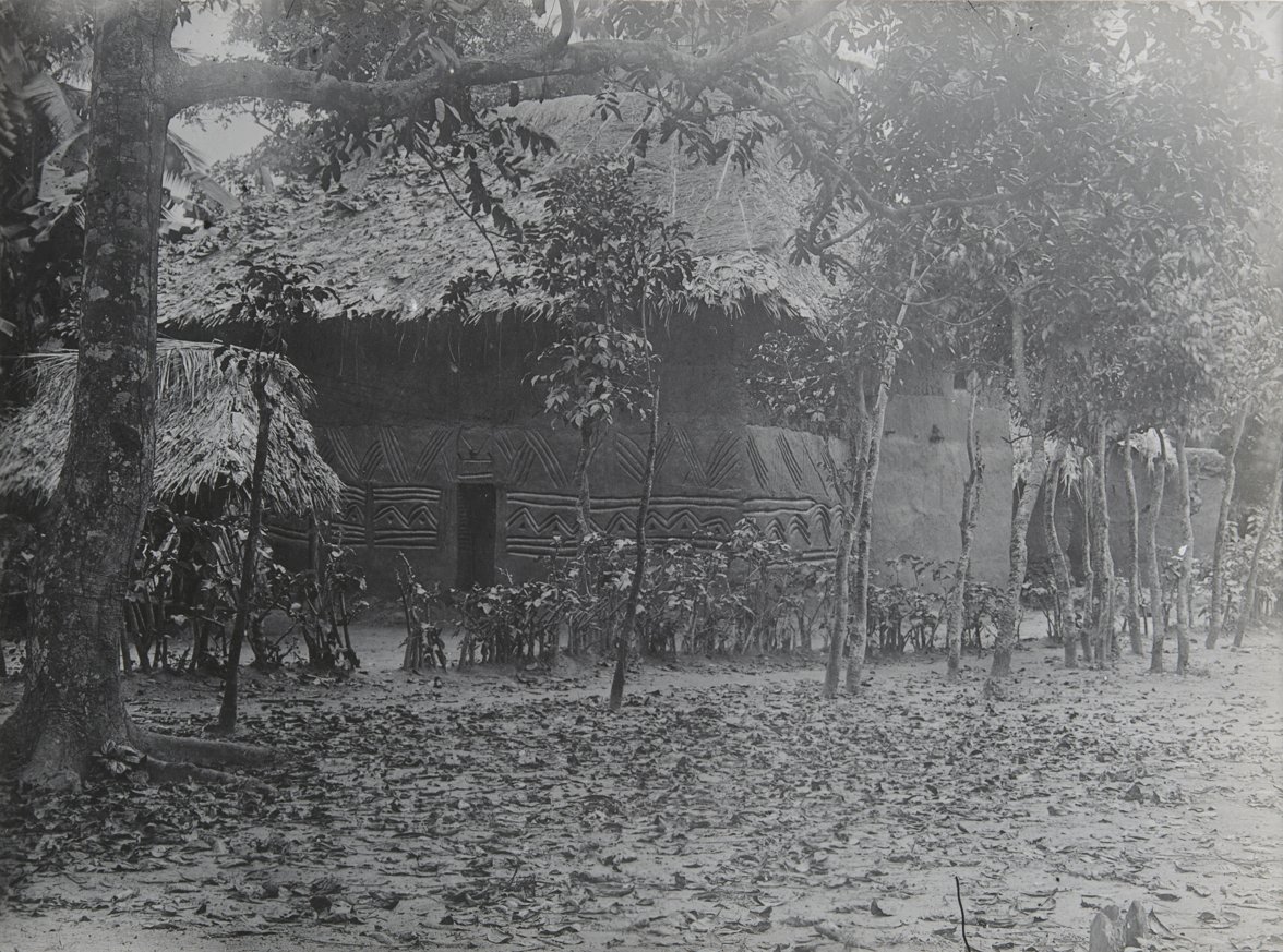 A building photographed in the western Igbo area, filed under Onicha Olona by the MAA Cambridge, but may be another surrounding Igbo town. The trees and shrubs appear to have been planted in an order. Photographed by Northcote Thomas and assistants, c. 1912-13.