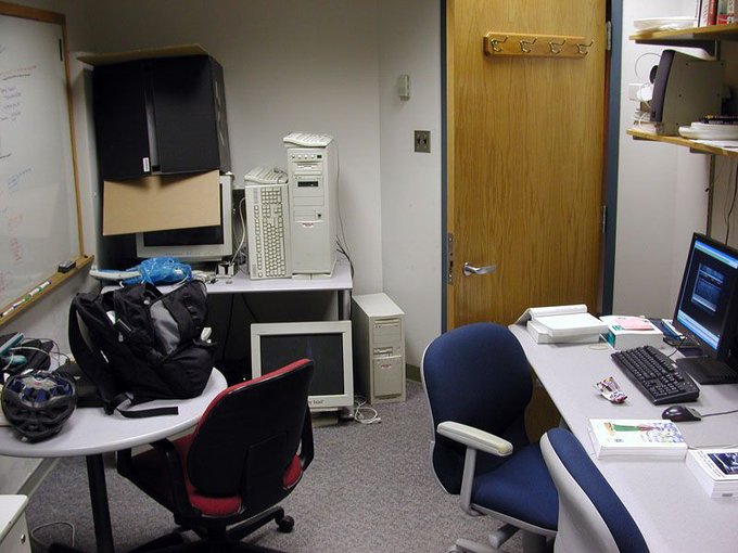 A photo of the interior of a small room in an office. On the right, there is a desk with a workstation and two swivel chairs. On the left, pieces of office furniture and disused computer equipment have been haphazardly stored against a wall with a marker board. There is gray low-pile carpet on the floor. The lighting is fluorescent.