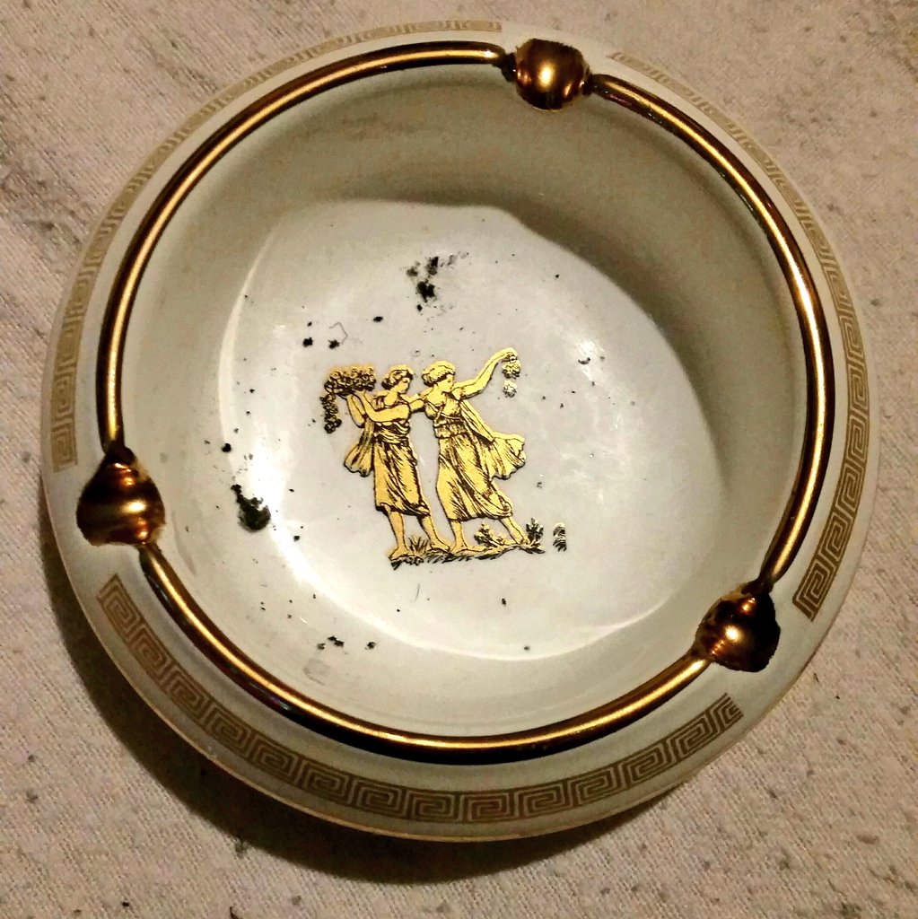 A round ceramic ashtray with greek-style gold decorations. A small nature scene of two women in ancient greek dress, one touching the other's shoulder fondly, decorates the bottom of the ashtray.