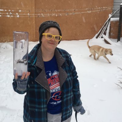 Snowy scene, a non-binary person in a blue jacket and gloves and yellow glasses holds up a rain gauge full of snow and a dog is seen running in the background under a clothesline