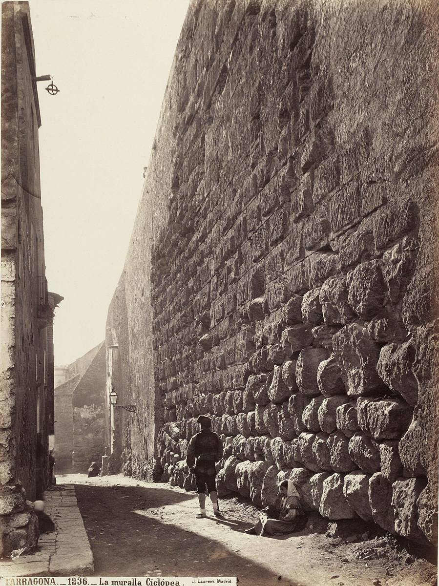 A young girl sits against the Roman cyclopean city wall of Tarragona while a man walks along its 2,000 year-old mortar free masonry, Spain ca. 1872