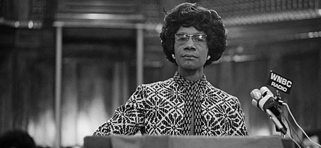 Image shows Shirley Chisholm staring into the camera, standing resolutely behind a podium with microphones.