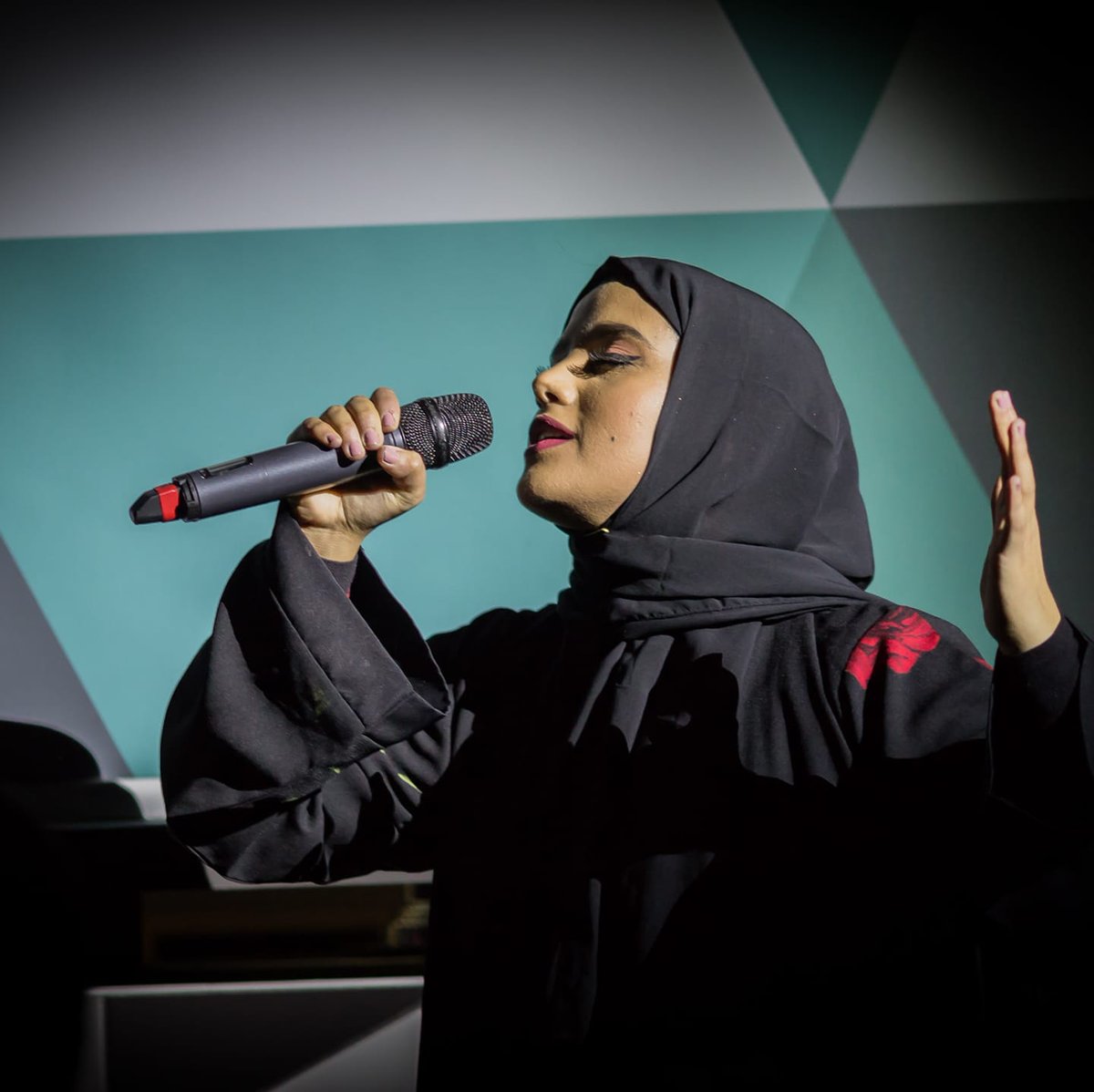 Amina Atik holds a microphone and appears to be singing into it/ She wears a black top and headscarf. 