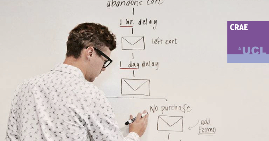 Male with glasses and black pen in his left hand standing in front of a whiteboard drawing a graph.