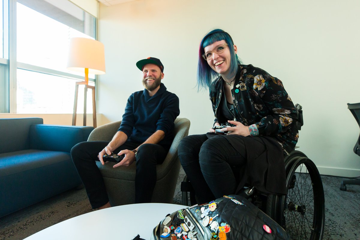 A bearded man and a woman in a wheelchair both smile as they play Xbox One together.