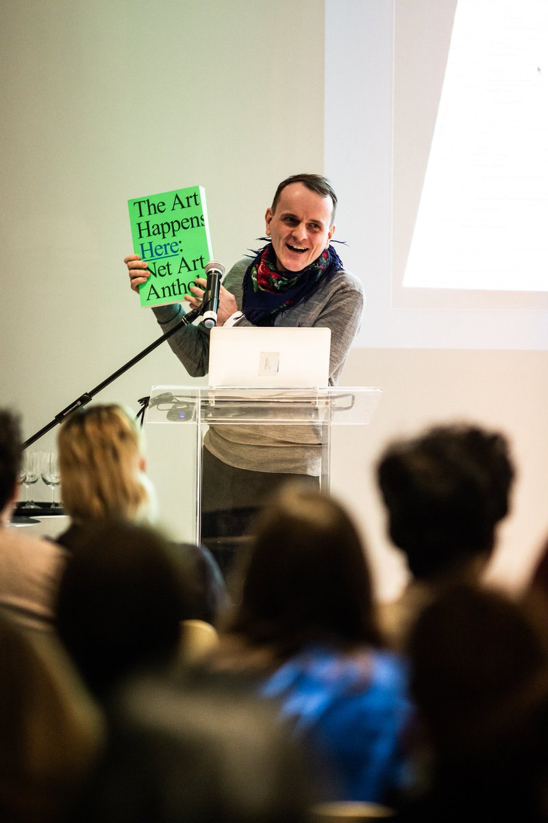 Dragan Espenschied stands at a lucite podium in front of a seated audience, holding a gorgeous green copy of 'the art happens here,' while smiling broadly.
