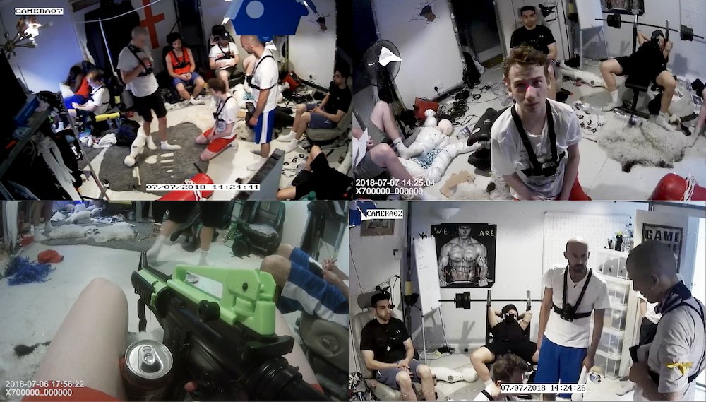 A still image from 'Cel' by Ed Fornieles. The screen is divided into four windows, offering CCTV-like views of a messy, nondescript apartment crowded with 20-something year old white men. They mostly wear white t-shirts, cotton shorts, and white cotton socks, which somehow makes them seem more like chavs.