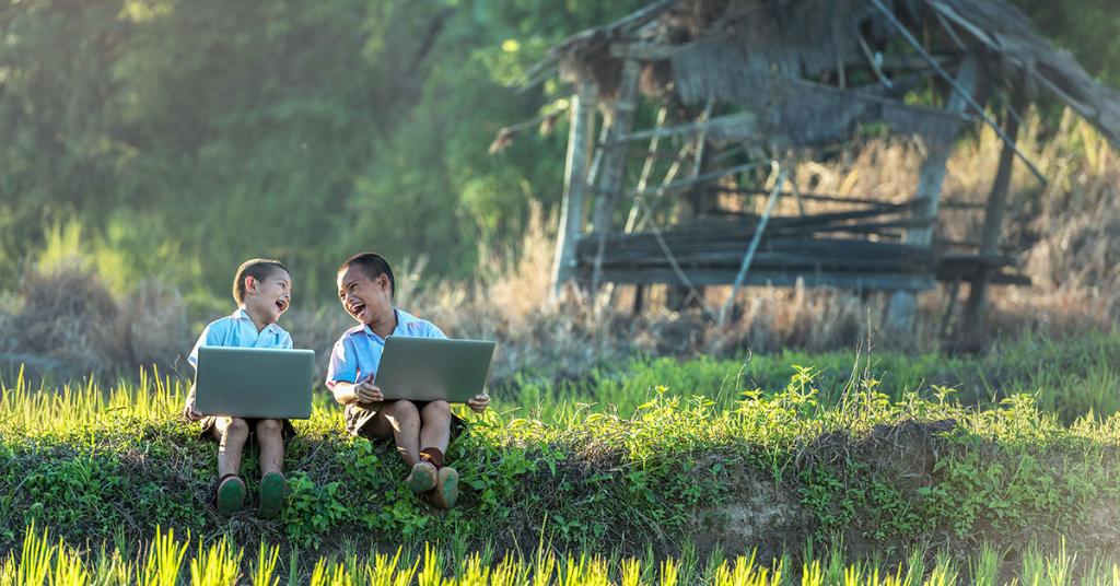 Photo of two young kids sitting with laptops laughing at one another.