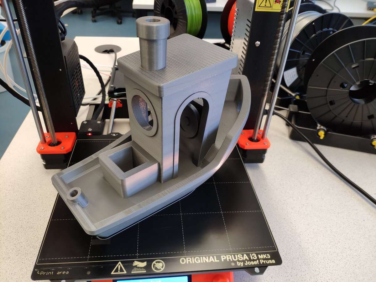 Ein Prusa 3D Drucker, darauf befindet sich ein, auf das maximale Druckvolumen hochskaliertes Modell des beliebten Benchmark-Bauteils "Benchy", welches ein comicartiges Boot ist.