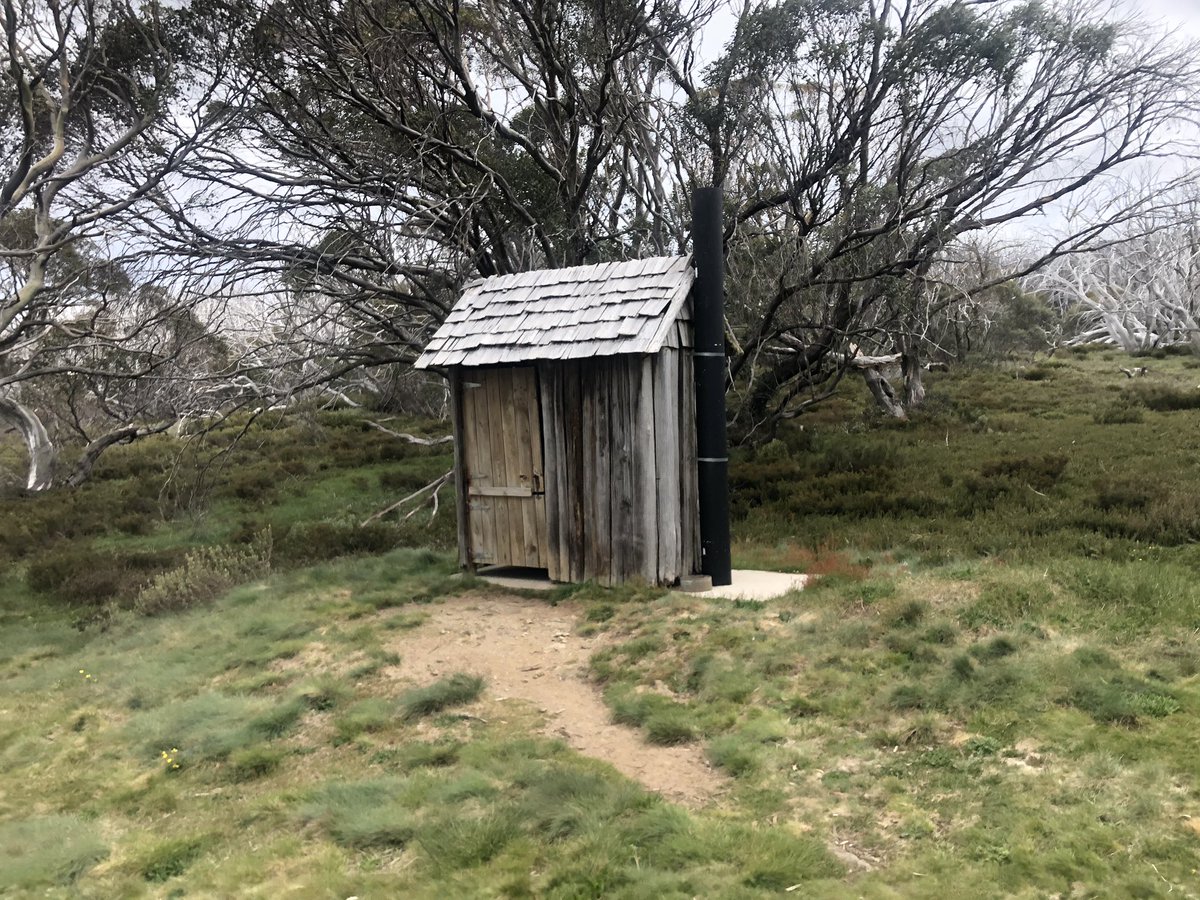 Classic Aussie Alpine hut Wallace’s Hut, the stylish outhouse, the snow patch in the gully up the hill and the view far below  #AAWT