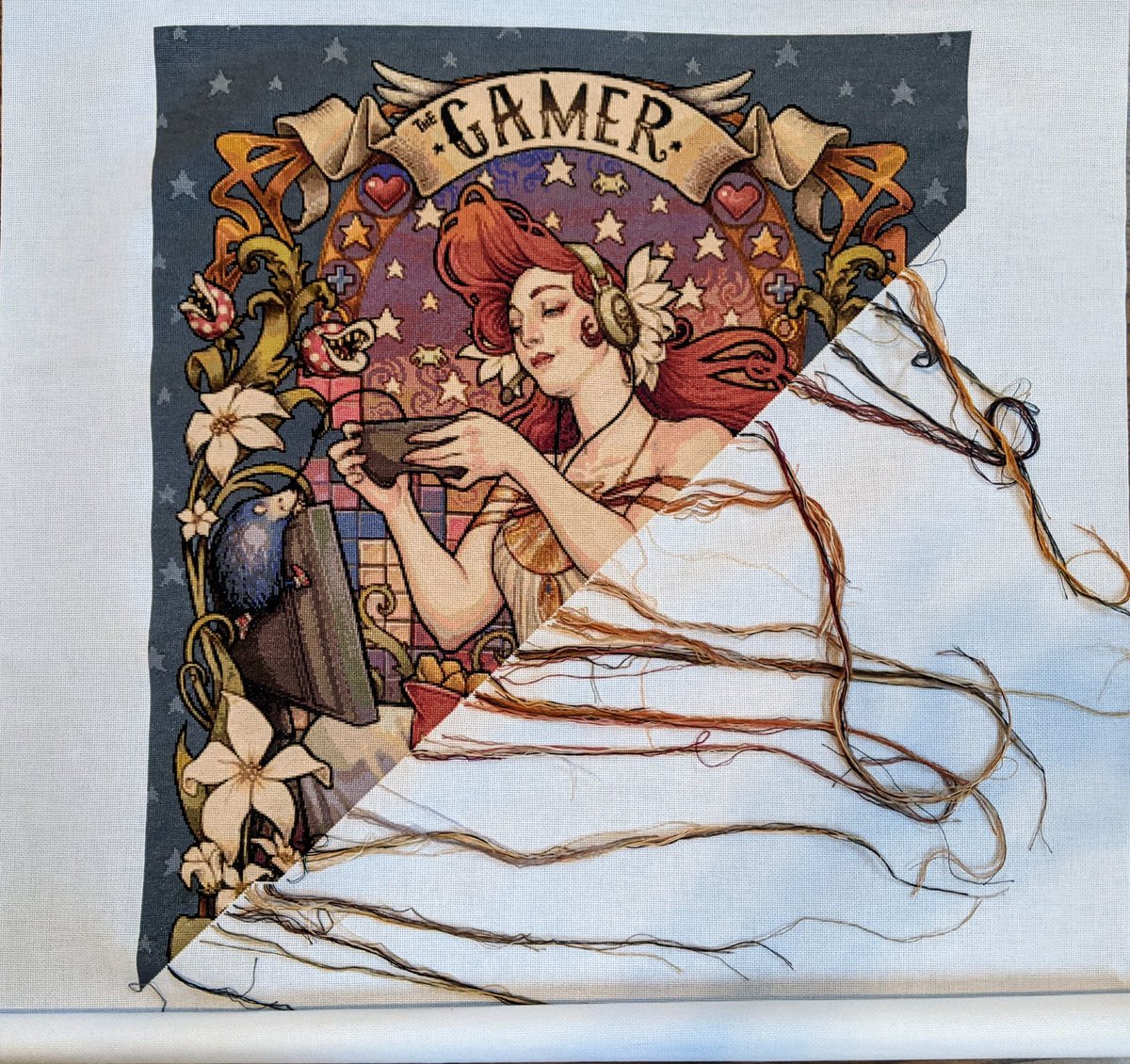 A poster-sized, partially completed cross stitch project showing a woman playing video games in art nouveau style.