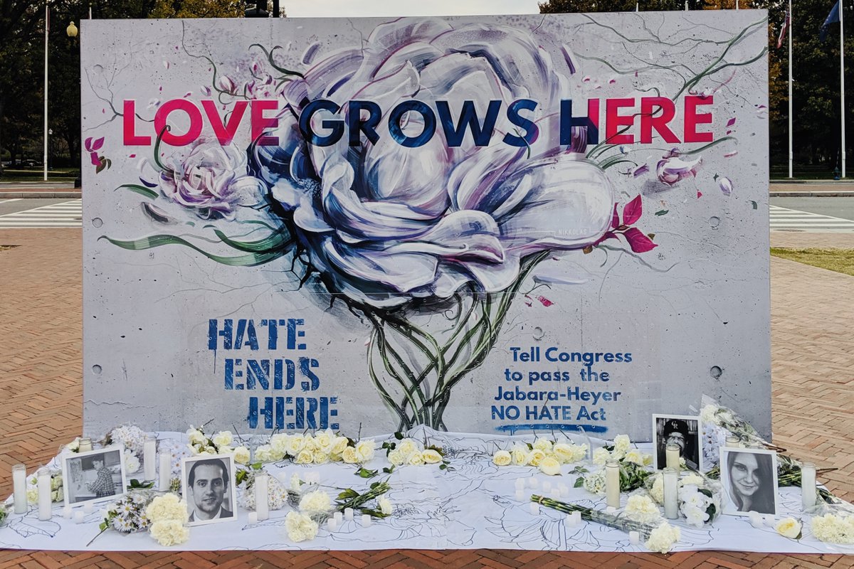 A mural outside Union Station in Washington, DC says "Love Grows Here" and "Hate Ends Here" and also reads "Tell Congress to pass the Jabara-Heyer NO HATE Act. Below the mural, candles, white flowers, and photographs of hate crime victims are displayed.