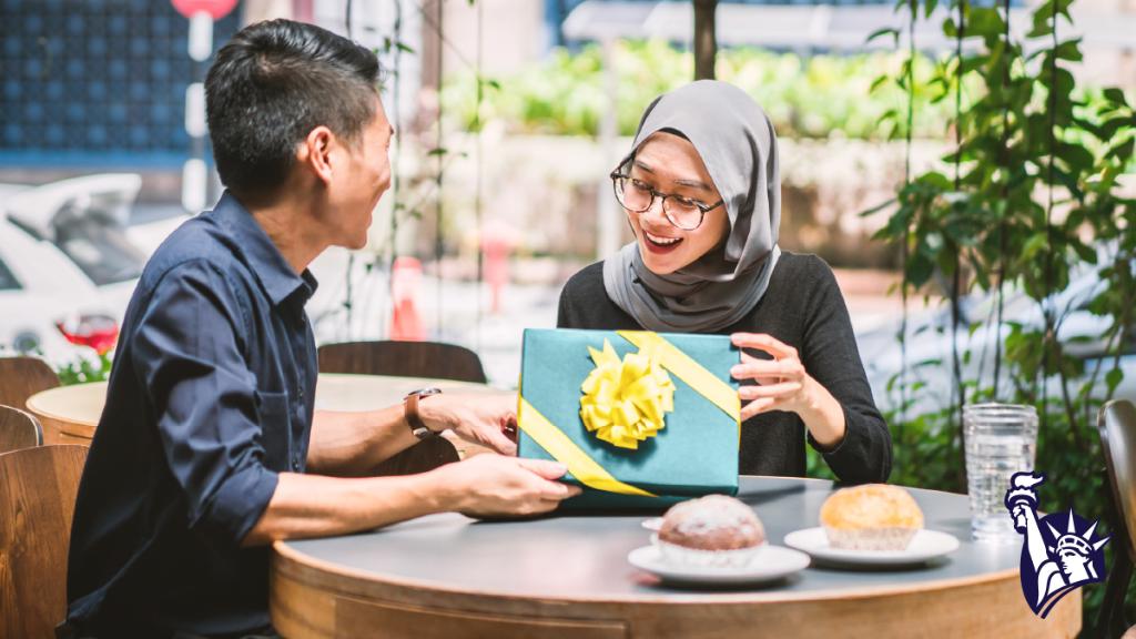A man wearing a dark blue button-down gives a gift wrapped in teal paper with a yellow ribbon to a girl wearing glasses, a black shirt, and a gray hijab. They sit at a round table with two muffins on white plates in front of them.