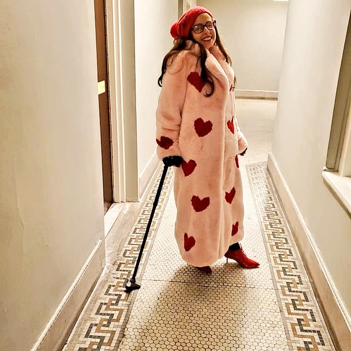 Smiling woman with long hair and red glasses wearing a red hat, pink furry Muppet coat with red hearts and red patent boots with kitten heels, and cane