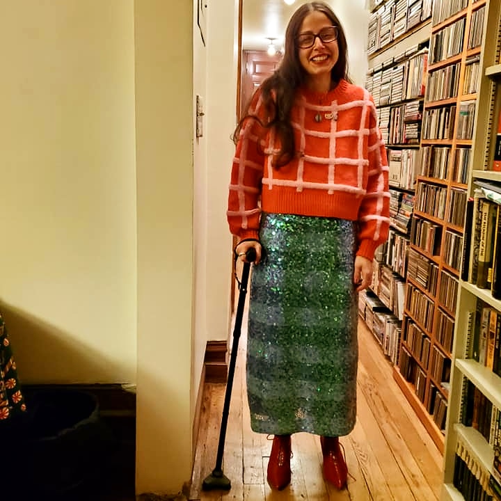 Smiling woman with long hair and red glasses wearing a red and pink sweater, mermaid color sequined skirt, red patent boots with kitten heels and a cane