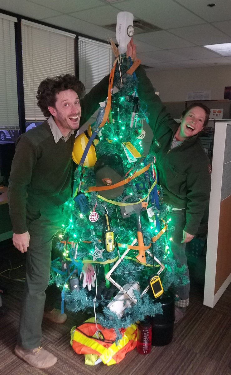 Two park rangers set a jug atop to a Christmas tree with green lights decorated with various research tools including a hatchet, hard hat, radio, NPS ball cap, sharpie marker, and more.
