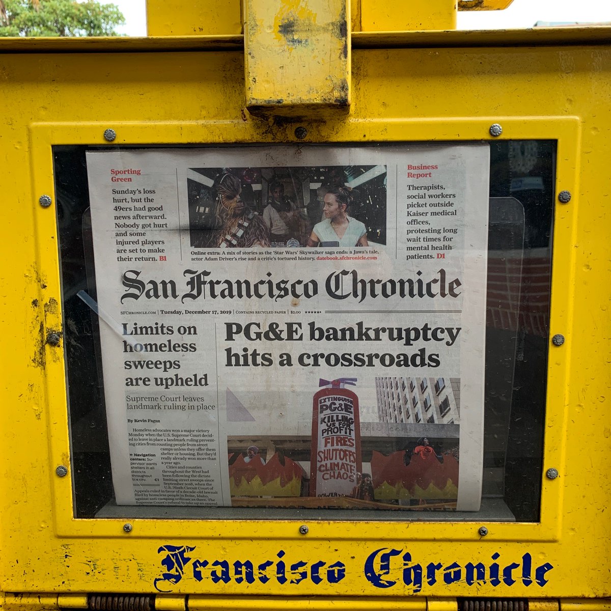SF chronicle in newspaper dispenser, front page story says "PG&E bankruptcy hits a crossroads" lead image is from yesterday's action, giant fire extinguisher and flames in front of pge headquarters