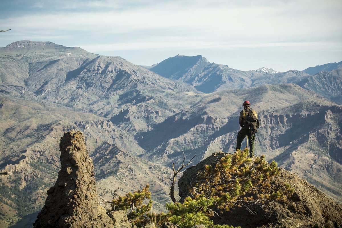A firefighter stands on a rocky outcrop above a large valley.
