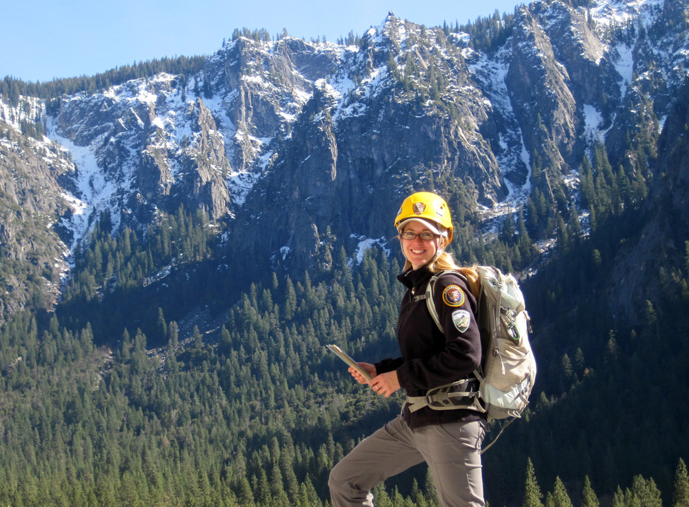 Girl wearing a hard hat and backpack stands in front of a snow covered mountain range